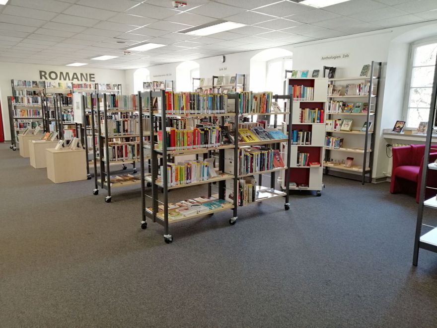 Das Bild zeigt die Romanabteilung der Bibliothek. Viele Regale mit Büchern und Sesseln zum ausruhen sind zu sehen.